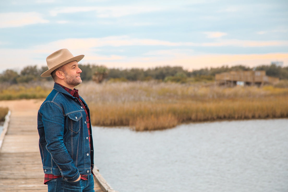 A man wearing a tan cowboy hat, blue denim jacket, and matching jeans stands on a wooden dock by a calm lake, looking out toward marshlands under a cloudy sky.